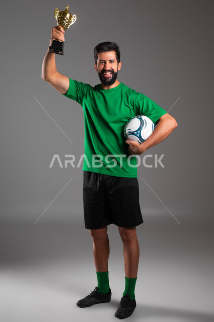 الشعور بالفرح والسرور، ممارسة الرياضات البدنية الممتعة، احتراف رياضة كرة القدم، النظر إلى الكاميرا بإيماءات الفوز والانتصار، بورتريه للاعب عربي سعودي خليجي مبتسم يرتدي الملابس الرياضية يحمل بيده الكرةويرفع كأس البطولة عاليا، صورة لكامل الجسم، خلفية رمادية .