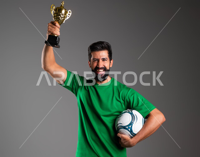 Feeling of joy and pleasure, professionalism in the sport of football, looking at the camera with gestures of victory and victory, a close-up portrait of a smiling Saudi Arabian Gulf player wearing sports clothing, holding the ball in his hand and raising the championship cup high, gray background