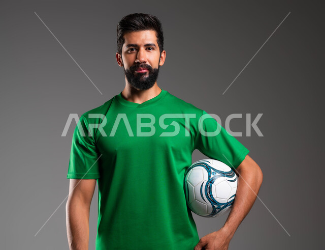 النظر إلى الكاميرا بإيماءات التفاؤل، الاستعداد لبدء المباراة، ممارسة هواية اللعب بكرة القدم، بورتريه مقرب للاعب عربي سعودي خليجي يرتدي الملابس الرياضية يحمل الكرة بيده، احتراف الرياضات الجماعية، خلفية رمادية .