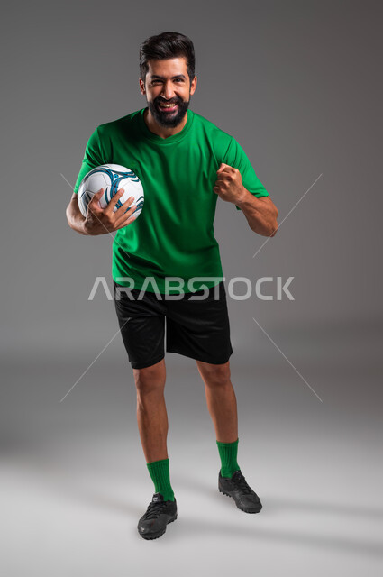 الشعور بالفرح والسرور، النظر إلى الكاميرا بإيماءات الفوز والانتصار، احتراف رياضة كرة القدم، بورتريه للاعب عربي سعودي خليجي مبتسم يرتدي الملابس الرياضية يرفع يده إلى الأعلى ويحمل بيده الكرة، صورة الجسم بالطول الكامل، خلفية رمادية .