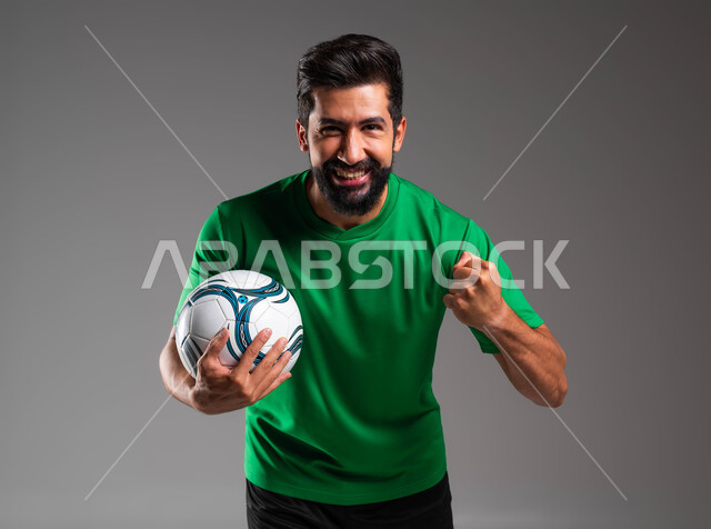النظر إلى الكاميرا بإيماءات الفوز والانتصار، ممارسة هواية اللعب بكرة القدم، احتراف الرياضات الجماعية، بورتريه مقرب للاعب عربي سعودي خليجي مبتسم يرتدي الملابس الرياضية يرفع يده إلى الأعلى ويحمل بيده الكرة، خلفية رمادية .
