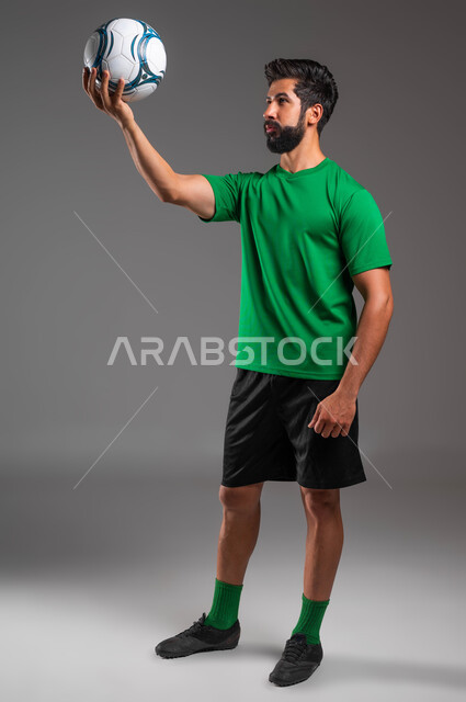 ممارسة هواية اللعب بكرة القدم، احتراف الرياضات الجماعية، إيماءات الانتباه والتركيز على الهدف، بورتريه مقرب للاعب عربي سعودي خليجي يرتدي الملابس الرياضية يقوم برفع الكرة إلى الأعلى وينظر إليها، صورة الجسم بالطول الكامل، خلفية رمادية .