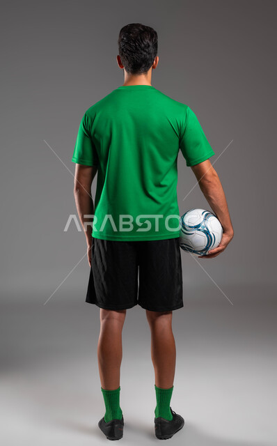 الوقوف باستقامة والاستعداد لبدء المباراة، ممارسة الرياضات الجماعية، بورتريه من الخلف للاعب عربي سعودي خليجي يرتدي الملابس الرياضية يحمل الكرة بيده، هواية اللعب بكرة القدم، صورة الجسم بالطول الكامل، خلفية رمادية .