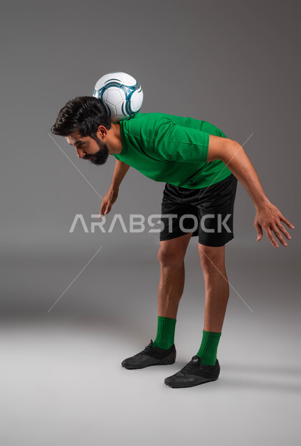 الحماس والاستعداد للمباراة، حركات التوازن والثبات، بورتريه مقرب للاعب عربي سعودي خليجي يرتدي الملابس الرياضية يضع كرة القدم على ظهره، فنون ومهارات اللعب، احترف التعامل مع الكرة، خلفية رمادية .