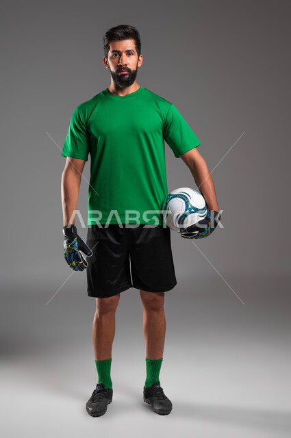 الوقوف باستقامة والاستعداد لبدء المباراة، ممارسة هواية اللعب بكرة القدم، بورتريه مقرب لحارس مرمى عربي سعودي خليجي يرتدي الملابس الرياضية وقفازات الوقاية ينظر إلى الكاميرا يحمل الكرة بيده، احتراف الرياضات الجماعية، صورة الجسم بالطول الكامل، خلفية رمادية .