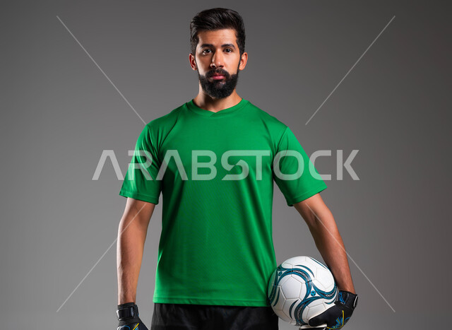 الاستعداد لبدء المباراة، الوقوف باستقامة والنظر إلى الكاميرا، ممارسة هواية اللعب بكرة القدم، بورتريه مقرب لحارس مرمى عربي سعودي خليجي يرتدي الملابس الرياضية وقفازات الوقاية يحمل الكرة بيده، احتراف الرياضات الجماعية، خلفية رمادية .