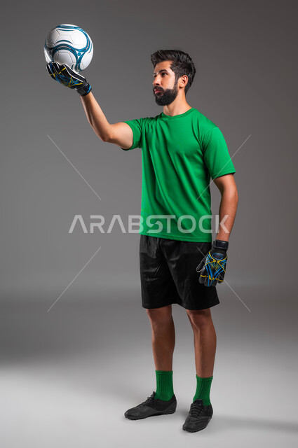 Standing straight and focusing on the goal, practicing the hobby of playing football, a close-up portrait of a Saudi-Gulf Arab goalkeeper wearing sports clothing and protective gloves, raising the ball up and looking at it, professional team sports, full-length body photo, gray background