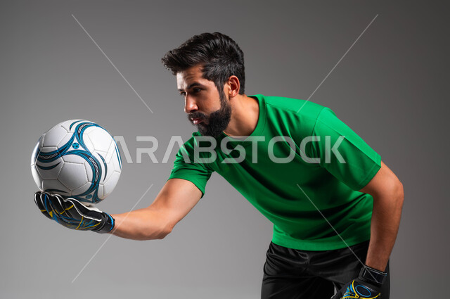 ممارسة هواية اللعب بكرة القدم، بورتريه مقرب لحارس مرمى عربي سعودي خليجي يرتدي الملابس الرياضية وقفازات الوقاية يحمل الكرة بكفه وينظر إليها، إيماءات الانحناء والتركيز على الهدف، احتراف الرياضات الجماعية، خلفية رمادية .