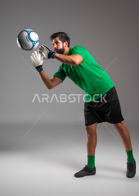 القفز والتصدي للأهداف، ممارسة رياضة كرة القدم، حارس مرمى يرتدي قفازات الوقاية يمنع دخول الهدف، إيماءات التركيز والدقة، بورتريه للاعب عربي سعودي خليجي يرتدي الملابس الرياضية يقوم بالتقاط الكرة، صورة الجسم بالطول الكامل، خلفية رمادية .