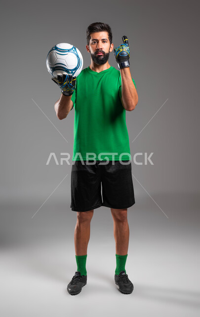 ممارسة رياضة كرة القدم، الحصول على المركز الاول، حارس مرمى يرتدي قفازات الوقاية يحمل الكرة بكفه، بورتريه للاعب عربي سعودي خليجي مبتسم يرتدي الملابس الرياضية ينظر إلى الكاميرا يرفع اصبع السبابة للأعلى، صورة الجسم بالطول الكامل، خلفية رمادية .