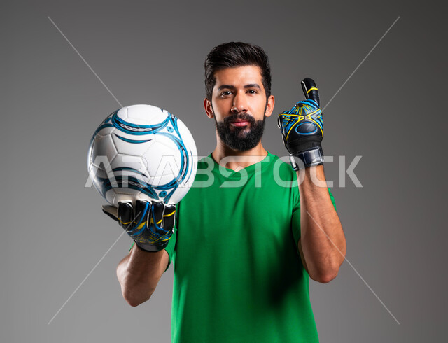 Obtaining first place, professional team sports, goalkeeper wearing protective gloves holding the ball in his palm, gestures of concentration and accuracy, portrait of a smiling Saudi Arabian Gulf player wearing sports clothing looking at the camera, raising his index finger up, gray background
