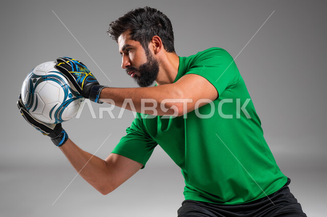 A goalkeeper wearing protective gloves prevents the goal from entering, attention, focus and accuracy, a close-up portrait of a Saudi-Gulf Arab player wearing sports clothing, picking up the ball in his hand, practicing football, professional team sports, gray background