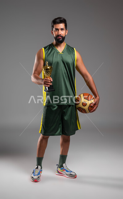 الفوز في فعاليات وتحديات بكرة السلة، المحافظة على أسلوب حياة صحي، احتراف الرياضات الجماعية، بورتريه لشاب عربي خليجي سعودي يرتدي الملابس الرياضية يمسك بيده الكرةوكأس البطولة، ممارسة التمرينات الرياضية، صورة الجسم بالكامل، خلفية رمادية.