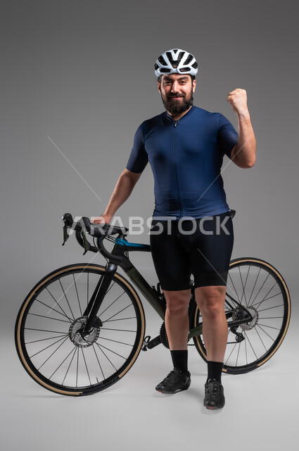 الاستعداد لقيادة ركوب الدراجة الهوائية، مسابقات ركوب الدراجات في السعودية، الاهتمام باللياقة البدنية، بورتريه لرجل عربي خليحي سعودي يرتدي الملابس الرياضية وخوذة الحماية ينظر إلى الكاميرا بحركات يد تدل على الحماس بالفوز، صورة لكامل الجسم، خلفية رمادية