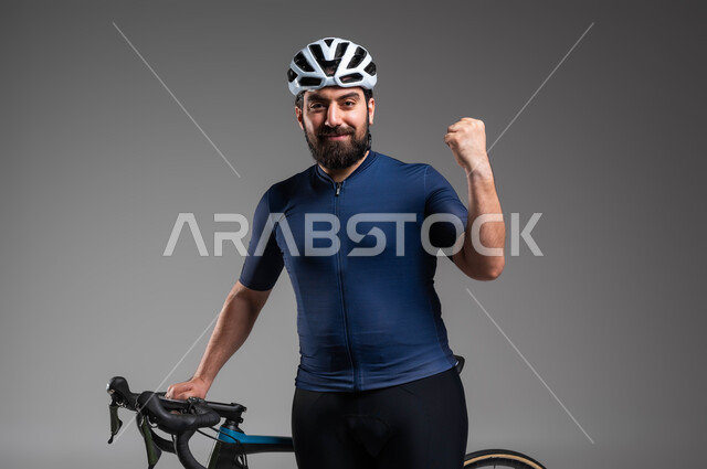 النظر إلى الكاميرا بحركات يد تدل على الحماس بالفوز، مسابقات ركوب الدراجات في السعودية، استخدام المواصلات الآمنة، الاهتمام باللياقة البدنية، بورتريه مقرب لرجل عربي خليحي سعودي يرتدي الملابس الرياضية وخوذة الحماية يستعد لقيادة دراجة السباق الهوائية، خلفية رمادية