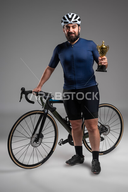 الحصول على كأس البطولة، إيماءات تدل على السعادة بالفوز في مسابقة ركوب الدراجة الهوائية في السعودية، العناية والاهتمام باللياقة البدنية، بورتريه لرجل عربي خليحي سعودي يرتدي الملابس الرياضية وخوذة الحماية، صورة لكامل الجسم، خلفية رمادية