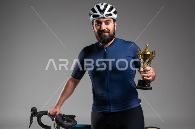 إيماءات تدل على السعادة بالفوز في مسابقة ركوب الدراجة الهوائية في السعودية، العناية والاهتمام باللياقة البدنية، بورتريه مقرب لرجل عربي خليحي سعودي يرتدي الملابس الرياضية وخوذة الحماية يحمل بيده كأس البطولة، خلفية رمادية
