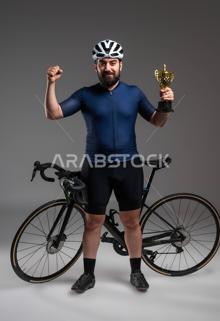 الحصول على كأس البطولة، إيماءات تدل على السعادة بالفوز في مسابقة ركوب الدراجة الهوائية في السعودية، العناية والاهتمام باللياقة البدنية، بورتريه لرجل عربي خليحي سعودي يرتدي الملابس الرياضية وخوذة الحماية، صورة لكامل الجسم، خلفية رمادية