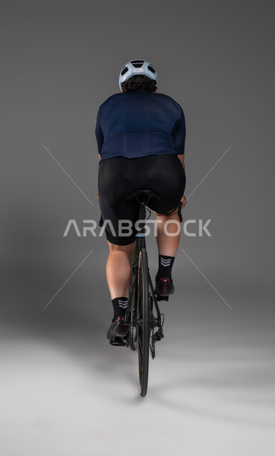 ممارسة الأنشطة البدنية الممتعة، فعاليات ركوب الدراجات في المملكة، مواصلات آمنة في السعودية، بورتريه من الخلف لرجل عربي خليحي سعودي يرتدي الملابس الرياضية وخوذة الحماية يقوم بقيادة دراجة السباق الهوائية، المحافظة على اللياقة الجسدية، خلفية رمادية