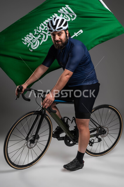 Raising the flag of the Kingdom of Saudi Arabia, practicing fun physical activities, cycling competitions and events, a portrait of a Saudi Gulf Arab man wearing sports clothing and a protective helmet riding a racing bicycle, maintaining physical fitness, gray background
