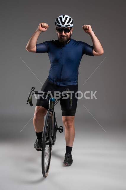 النظر إلى الكاميرا بحركات يدين تدل على الحماس بالفوز، مسابقات ركوب الدراجات في السعودية، استخدام المواصلات الآمنة، الاهتمام باللياقة البدنية، بورتريه لرجل عربي خليحي سعودي يرتدي الملابس الرياضية وخوذة الحماية يستعد لقيادة دراجة السباق الهوائية، خلفية رمادية