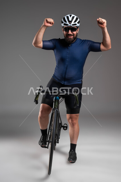 النظر إلى الكاميرا بحركات يدين تدل على الحماس بالفوز، مسابقات ركوب الدراجات في السعودية، استخدام المواصلات الآمنة، الاهتمام باللياقة البدنية، بورتريه لرجل عربي خليحي سعودي يرتدي الملابس الرياضية وخوذة الحماية يستعد لقيادة دراجة السباق الهوائية، خلفية رمادية