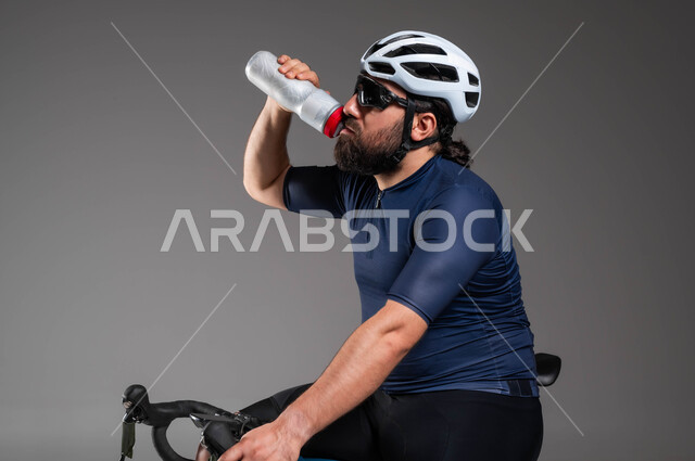 Using safe transportation, paying attention to physical fitness, cycling events in the Kingdom, practicing enjoyable physical activities, close-up portrait of a Saudi Gulf Arab man wearing sports clothing and a protective helmet, riding a racing bicycle and drinking water, gray background