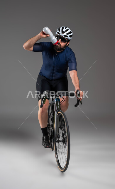 Using safe transportation, paying attention to physical fitness, cycling events in the Kingdom, practicing fun physical activities, a portrait of a Saudi Gulf Arab man wearing sports clothes and a protective helmet, riding a racing bicycle and drinking water, gray background.