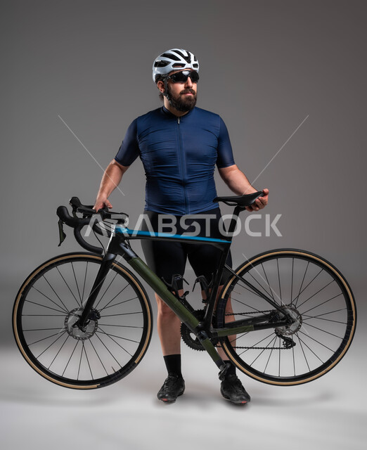 Safe transportation in Saudi Arabia, practicing fun physical activities, cycling events in the Kingdom, portrait of a Saudi Gulf Arab man wearing sports clothing and a protective helmet carrying a racing bicycle, maintaining physical fitness, gray background