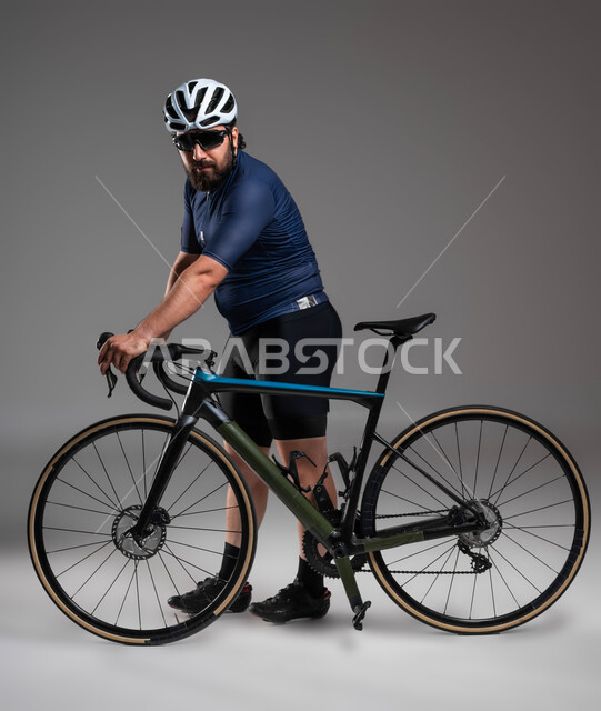 ممارسة الأنشطة البدنية الممتعة، مواصلات آمنة فيي السعودية، بورتريه لرجل عربي خليحي سعودي يرتدي الملابس الرياضية وخوذة الحماية يستعد لركوب دراجة السباق الهوائية، المحافظة على اللياقة الجسدية، صورة الجسم بالطول الكامل، خلفية رمادية