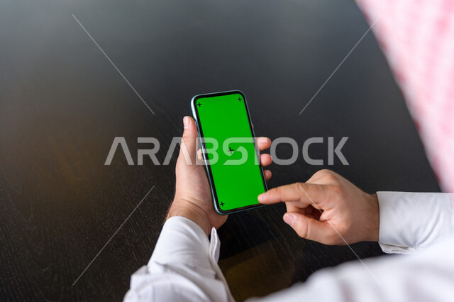 رجل أعمال سعودي خليجي يستخدام هاتفه المحمول بخلفية خضراء كروما ، تقنية و حاسب ، استخدام تقنية السمارت على الهاتف المحمول ليتحكم بالتلفاز