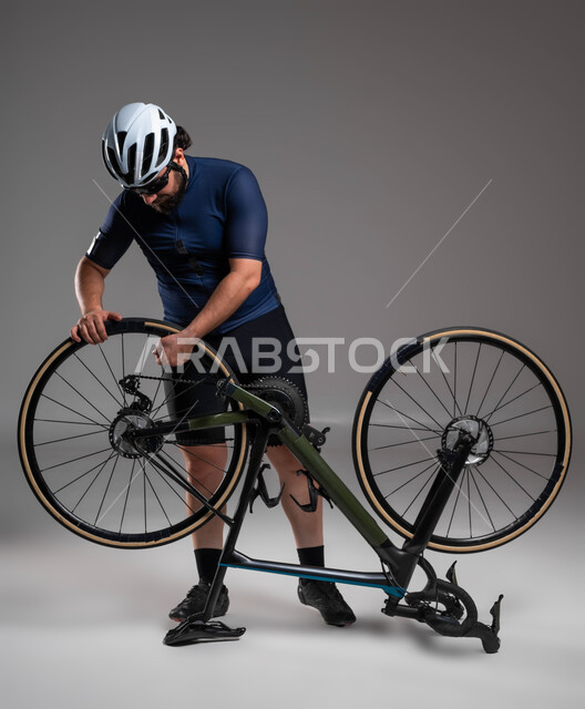 التعرض لحوادث وأعطال، ممارسة الأنشطة البدنية الممتعة، مسابقات وفعاليات ركوب الدراجات، بورتريه لرجل عربي خليحي سعودي يرتدي الملابس الرياضية وخوذة الحماية يقوم بإصلاح دراجة السباق الهوائية، المحافظة على اللياقة الجسدية، خلفية رمادية