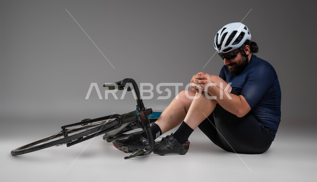 التعرض لحوادث وإصابات جسدية أثناء قيادة الدراجة الهوائية، مسابقات وفعاليات ركوب الدراجات، بورتريه لرجل عربي خليحي سعودي يرتدي الملابس الرياضية وخوذة الحماية يجلس على الأرض بإيماءات الألم، خلفية رمادية