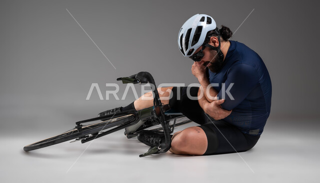 Physical pain and injuries, accidents while riding a bicycle, cycling competitions and events, portrait of a Saudi Khaleeji Arab man wearing sports clothes and a protective helmet sitting on the ground with pain gestures, gray background