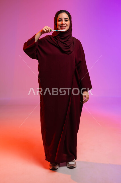 روتين نظافة الفم اليومية، تجنب التسوس وأمراض اللثة، النظر إلى الكاميرا بإيماءات السرور، بورتريه لشابة عربية خليجية سعودية تمسك بيدها فرشاة أسنانها، مفهوم العناية والاهتمام بصحة ونظافة الأسنان، صورة لكامل الجسم، خلفية ملونة