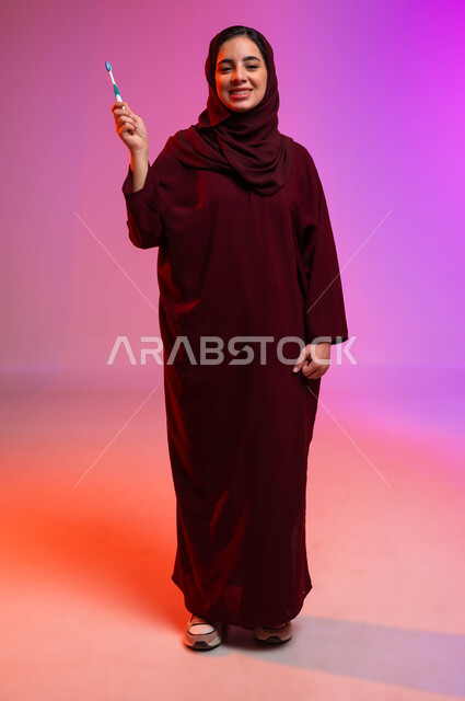 روتين نظافة الفم اليومية، تجنب التسوس وأمراض اللثة، النظر إلى الكاميرا بإيماءات السرور، بورتريه لشابة عربية خليجية سعودية تمسك بيدها فرشاة أسنانها، مفهوم العناية والاهتمام بصحة ونظافة الأسنان، صورة لكامل الجسم، خلفية ملونة