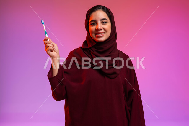 تجنب التسوس وأمراض اللثة، روتين نظافة الفم اليومية، النظر إلى الكاميرا بإيماءات السرور، بورتريه مقرب لشابة عربية خليجية سعودية تمسك بيدها فرشاة أسنانها، مفهوم العناية والاهتمام بصحة ونظافة الأسنان، خلفية ملونة