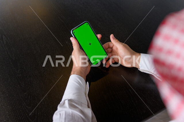 رجل أعمال سعودي خليجي يستخدام هاتفه المحمول بخلفية خضراء كروما ، تقنية و حاسب ، استخدام تقنية السمارت على الهاتف المحمول ليتحكم بالتلفاز