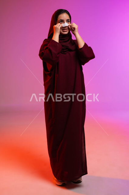 استخدام ماسكات تنظيف المسام ومكونات تجميلية نسائية، العناية والاهتمام بنضارة وتحسين البشرة، التخلص من الرؤوس السوداء والجلد الميت، بورتريه لشابة عربية خليجية سعودية محجبة ترتدي العباءة تقوم بوضع لصقات الأنف، صورة لكامل الجسم، خلفية ملونة