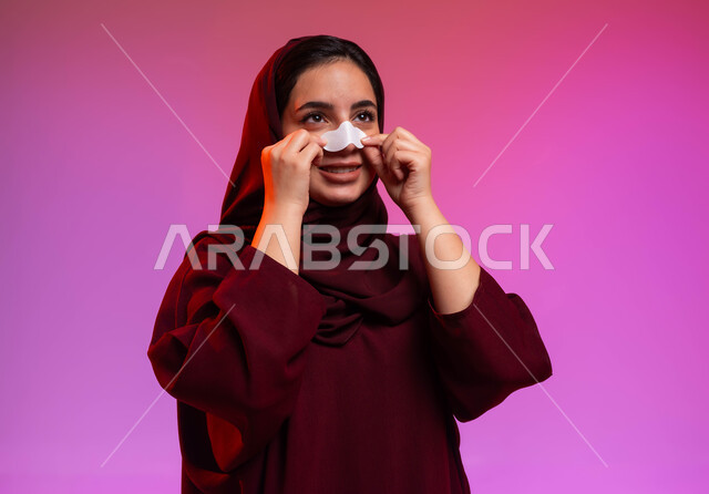 العناية والاهتمام بنضارة وتحسين البشرة، استخدام ماسكات تنظيف المسام ومكونات تجميلية نسائية، التخلص من الرؤوس السوداء والجلد الميت، بورتريه مقرب لامرأة عربية خليجية سعودية محجبة ترتدي العباءة تقوم بوضع لصقات الأنف، خلفية ملونة