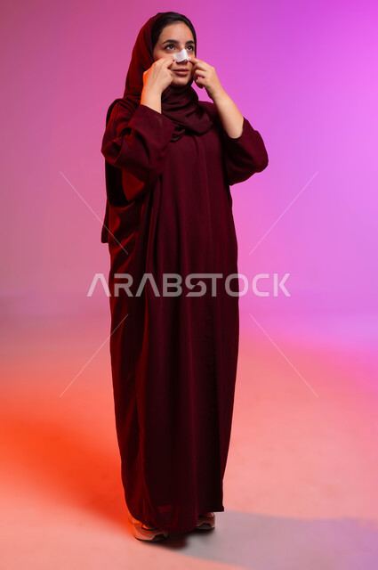 استخدام ماسكات تنظيف المسام ومكونات تجميلية نسائية، العناية والاهتمام بنضارة وتحسين البشرة، التخلص من الرؤوس السوداء والجلد الميت، بورتريه لامرأة عربية خليجية سعودية محجبة ترتدي العباءة تقوم بوضع لصقات الأنف، صورة لكامل الجسم، خلفية ملونة