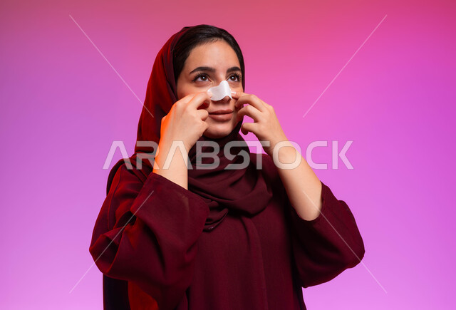العناية والاهتمام بنضارة وتحسين البشرة، استخدام ماسكات تنظيف المسام ومكونات تجميلية نسائية، التخلص من الرؤوس السوداء والجلد الميت، بورتريه مقرب لامرأة عربية خليجية سعودية محجبة ترتدي العباءة تقوم بوضع لصقات الأنف، خلفية ملونة