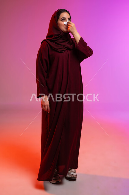 استخدام ماسكات تنظيف المسام ومكونات تجميلية نسائية، العناية والاهتمام بنضارة وتحسين البشرة، التخلص من الرؤوس السوداء والجلد الميت، بورتريه لامرأة عربية خليجية سعودية محجبة ترتدي العباءة تقوم بوضع لصقات الأنف، صورة لكامل الجسم، خلفية ملونة