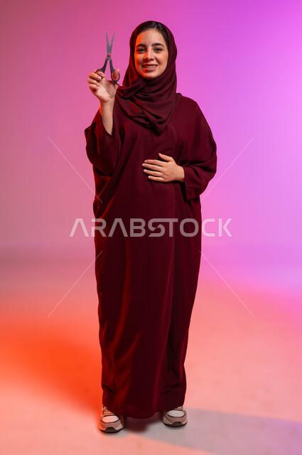 النظر إلى الكاميرا بإيماءات السرور، مركز تجميلي نسائي، مفهوم العناية و الاهتمام بالجمال والمظهر الخارجي، قص و تصفيف الشعر في صالون الحلاقة، بورتريه لامرأة عربية خليجية سعودية ترتدي العباءة والحجاب تحمل بيدها المقص، صورة الجسم بالكامل، خلفية ملونة.