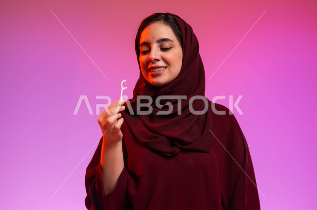 الاهتمام بالأناقة والمظهر الخارجي، مفهوم العناية بصحة الفم، التأكد من نظافة الأسنان بعد تناول الطعام، المحافظة على ابتسامة بيضاء، بورتريه مقرب لامرأة عربية خليجية سعودية تمسك بيدها الخيط السني، خلفية ملونة
