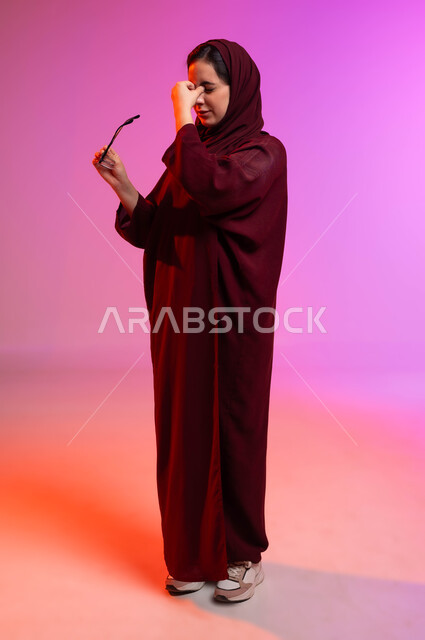 تحسين وتعديل الرؤية، المحافظة على صحة العينين، ارتداء النظارات الخاصة بضعف النظر، بورتريه من الجانب لسيدة عربية خليجية سعودية محجبة ترتدي العباءة تمسك النظاره الطبيه وتضع يدها على وجهها بإيماءات التعب والارهاق، صورة لكامل الجسم، خلفية ملونة