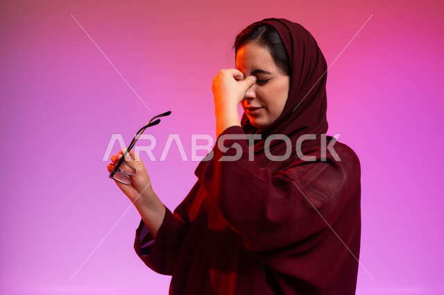 مفهوم تحسين وتعديل الرؤية، المحافظة على صحة العينين، ارتداء النظارات الخاصة بضعف النظر، بورتريه مقرب من الجانب لسيدة عربية خليجية سعودية محجبة ترتدي العباءة تمسك النظاره الطبيه وتضع يدها على وجهها بإيماءات التعب والارهاق، خلفية ملونة