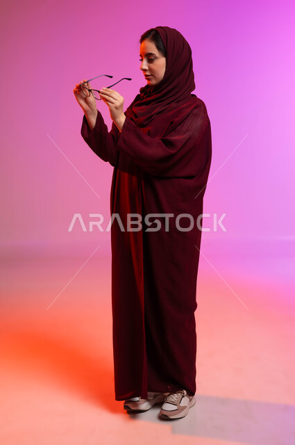 المحافظة على صحة العينين، ارتداء النظارات الخاصة بضعف النظر، مفهوم تحسين وتعديل الرؤية، بورتريه من الجانب لسيدة عربية خليجية سعودية محجبة ترتدي العباءة تمسك بيدها النظاره الطبيه، صورة الجسم بالطول الكامل، خلفية ملونة