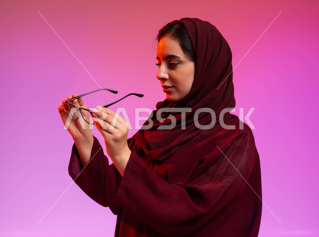 Maintaining eye health, wearing glasses for low vision, the concept of improving and modifying vision, a close-up portrait from the side of a veiled Saudi Arabian Gulf woman wearing an abaya, holding her prescription glasses in her hand, colored background