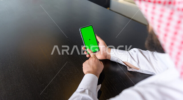 رجل أعمال سعودي خليجي يستخدام هاتفه المحمول بخلفية خضراء كروما ، تقنية و حاسب ، استخدام تقنية السمارت على الهاتف المحمول ليتحكم بالتلفاز